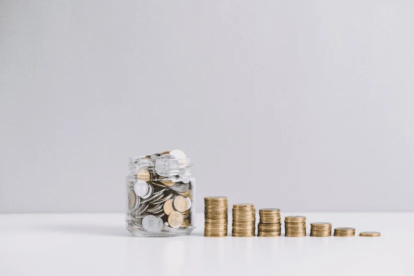 A glass jar filled with coins next to stacks of coins.