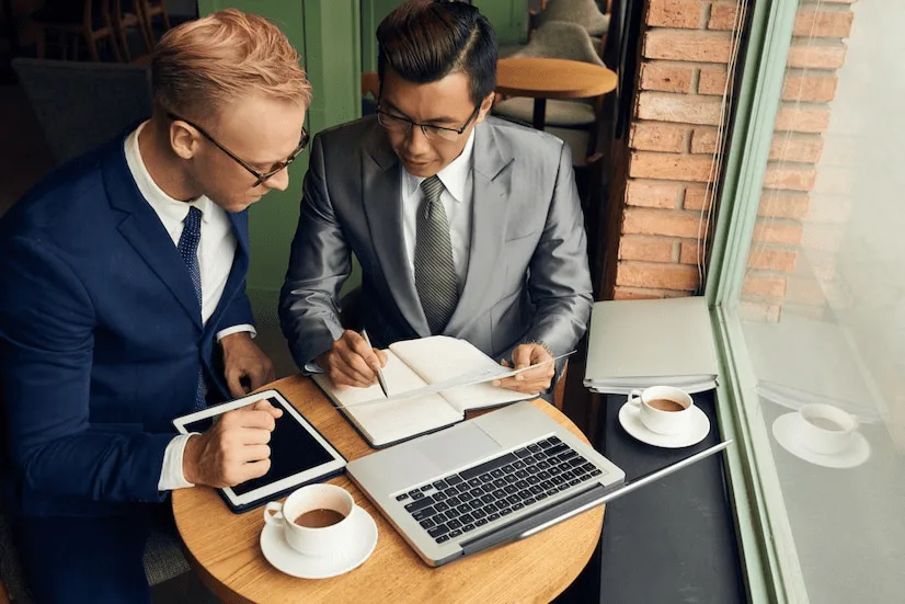 Two men sitting at a table with laptops and coffee.